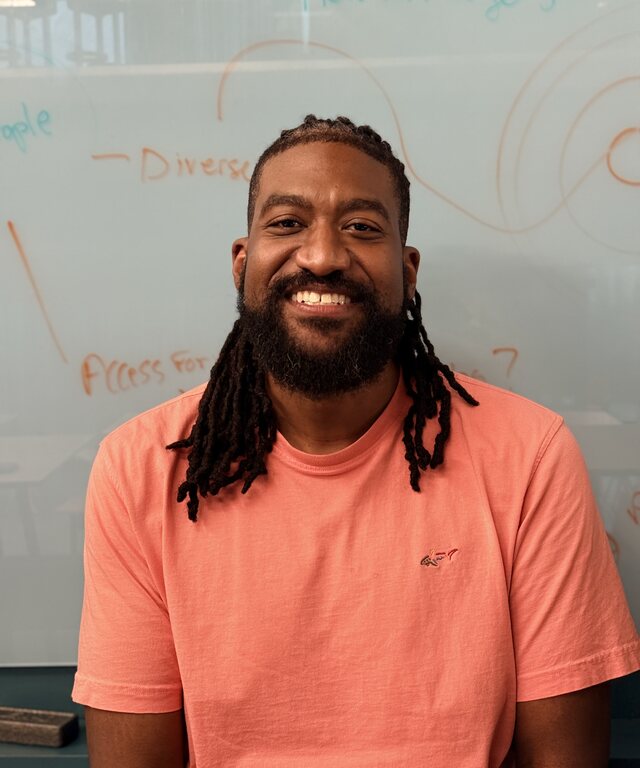 Mark Sanders, smiling, in a coral shirt in front of a whiteboard