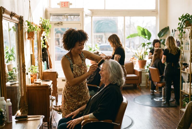 A stylist finishing an older woman's hair as she laughs at the mirror (an illustration)